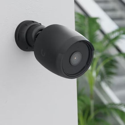 Caméra de vidéosurveillance noire fixée en applique sur un mur clair, orientée vers une baie vitrée et un jardin.